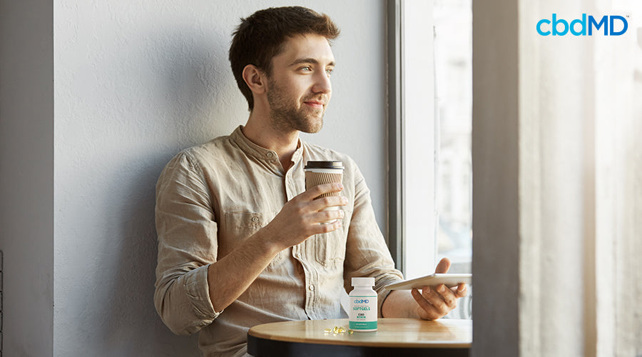 Um homem sentado com um café e um comprimido nas mãos ao lado de cápsulas de amplo espectro - cbdMD