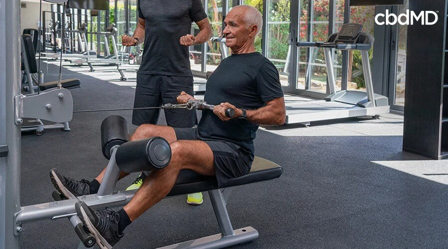 Un entrenador en un gimnasio mostrando a alguien cómo realizar un ejercicio en una máquina de ejercicios - cbdMD