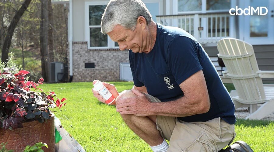 A man outside kneeling on the ground holding his knee with recover cream in his hand - cbdMD