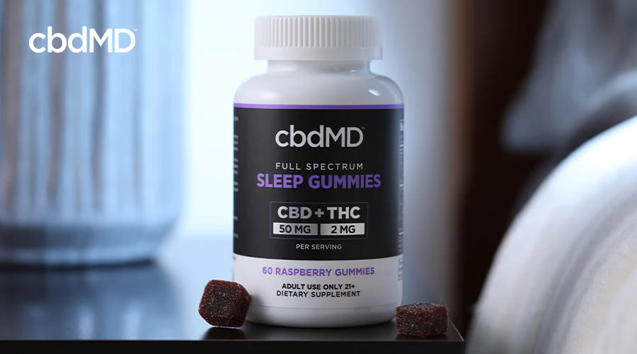 cbdMD Full Spectrum melatonin sleep aid gummies Recipiente de 1500 mg de framboesa com gomas ao lado em uma mesa de cabeceira.