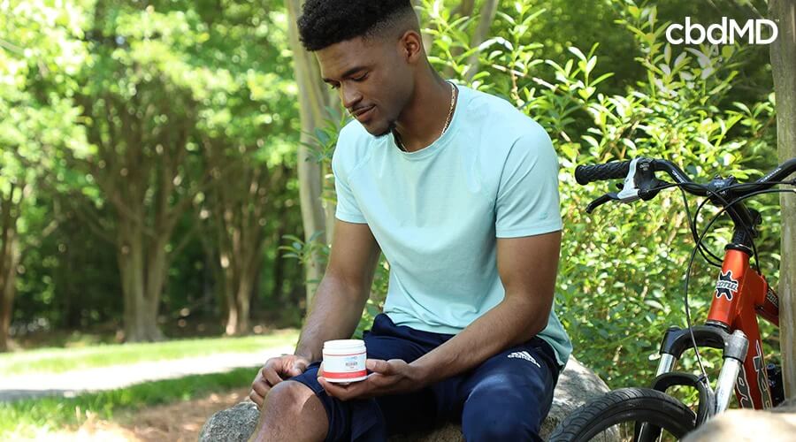 A man sitting on a rock outside next to his bike as he holds and uses premium recover cream - cbdMD