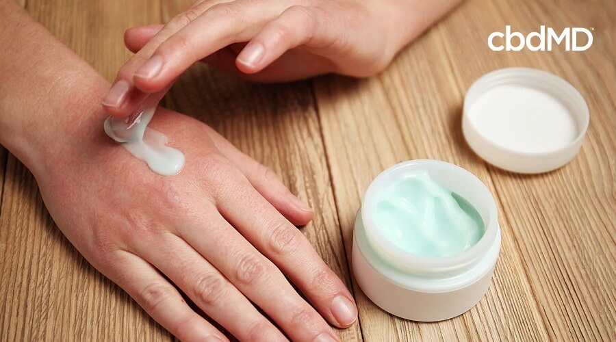 A feminine hand rubs light green cbd cream on the top as it rests on a wooden table
