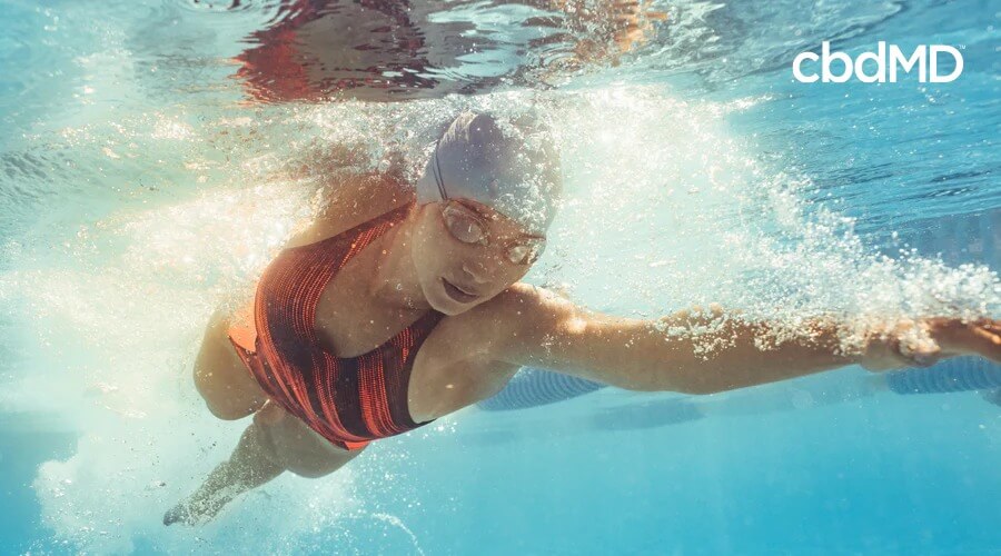 Uma mulher de maiô preto e laranja, touca de natação e óculos nada debaixo d'água em uma piscina