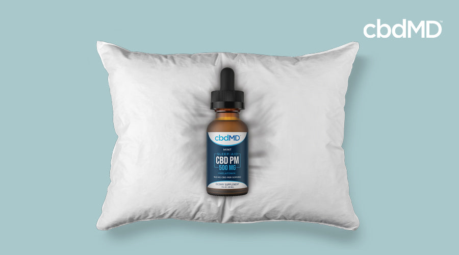 Tincture bottle of 500 mg CBD PM by cbdMD rests on white pillow with light blue background