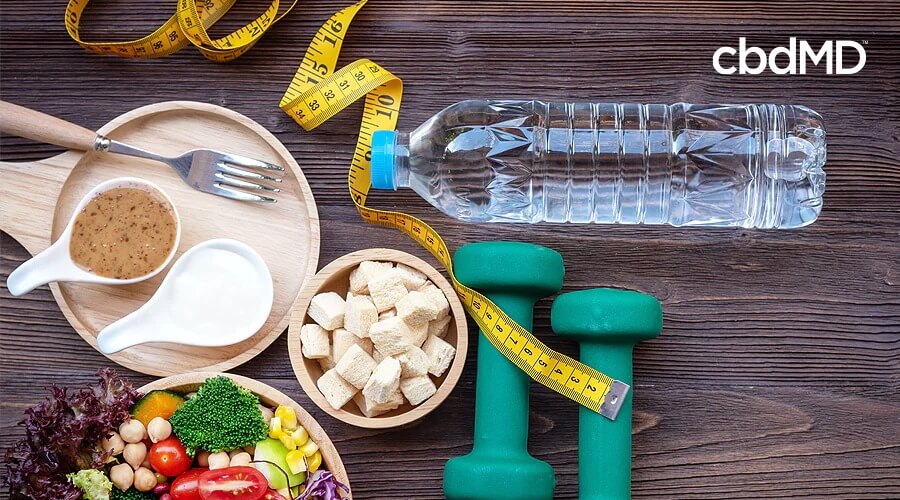 An assortment of healthy snacks sits along side a pair of weights, a water bottle, and a tape measure on a table