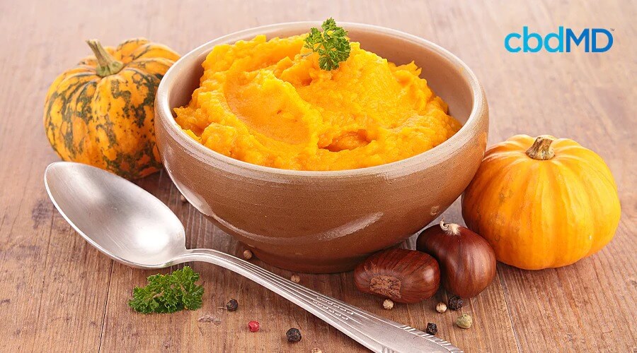A bowl of mashed pumpkin sits with a spoon beside it on a table near whole pumpkins
