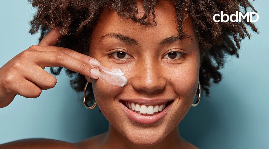 An attractive woman with dark skin and curly hair spreads cbd cream on her cheek