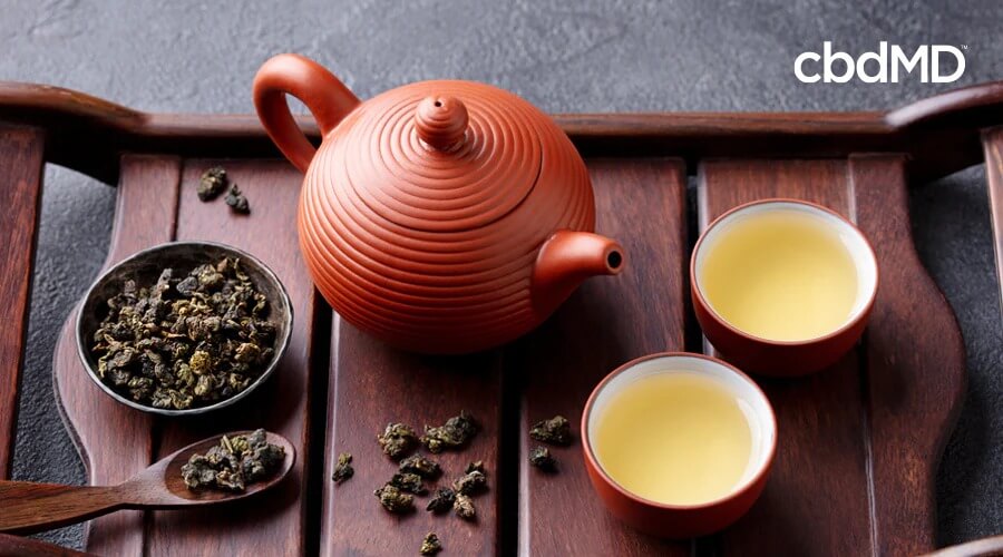 Dried tea leaves sit in a bowl next to a red teapot and two cups full of tea