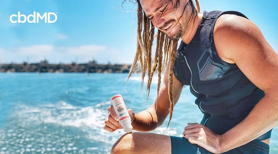 A man with dreadlocks on a surf board rubs cbd freeze topical onto his knee