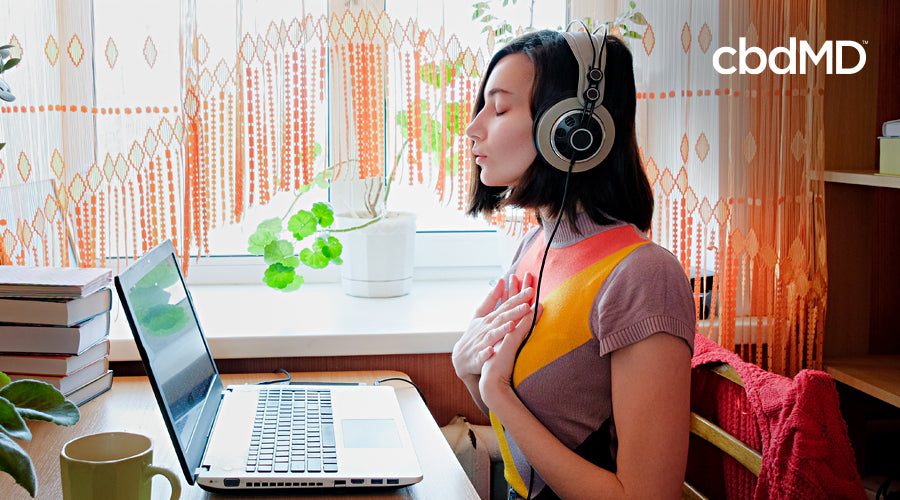 Mulher sentada à mesa com fones de ouvido e laptop cercada por plantas e livros com as mãos apoiadas no peito