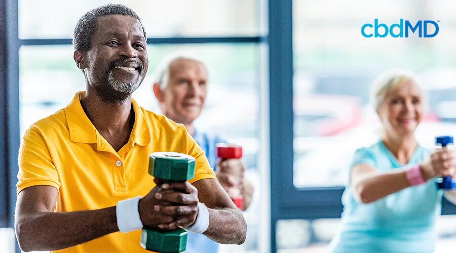 A group of older people hold weights as they workout and a dark skinned man with white hair stands in the front with a yellow shirt