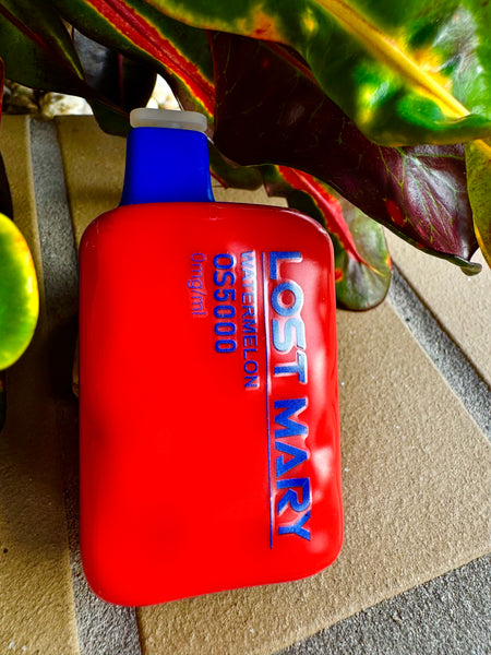 The image shows a bright red disposable vape device with a blue mouthpiece, lying on a tan surface. The side of the device is embossed with the words "LOST MARY WATERMELON 05G5000 0mg/ml" in white lettering, indicating the flavor and nicotine content. The device is positioned in front of a colorful backdrop of plant leaves with red, green, and yellow hues, which creates a dynamic contrast with the vivid red of the vape.