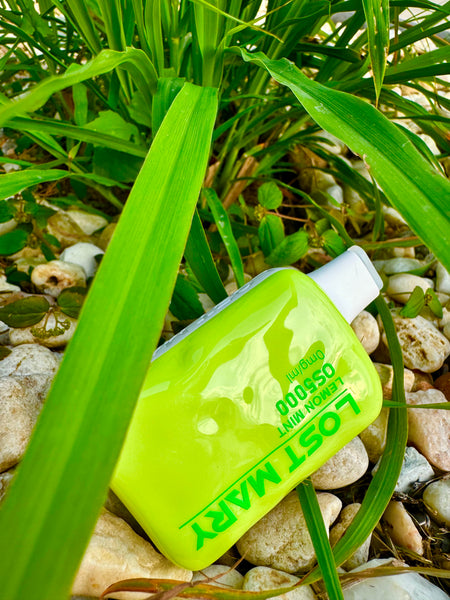 The image displays a neon green disposable vape device lying on a bed of white stones, partially covered by green grass and plant leaves. The device is oriented with the mouthpiece pointing downwards and the body of the vape angled upwards, showing the label "LOST MARY" in reverse, suggesting the photo has been taken from above with the vape device upside down. The text also includes "OS5000 0mg/ml", indicating the device's specifications. The natural greenery enveloping the device creates a dynamic contrast with its bright, artificial color.