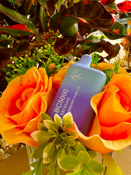 A gradient blue to purple disposable vape device labeled "BC5000 EBCREATE Blueberry Pomegranate" is tucked into a colorful bouquet featuring artificial yellow-orange roses. The device is positioned vertically, with its white mouthpiece peeking out from the top of the bouquet. The surrounding silk flowers and green leaves add vibrant color and a touch of lifelike texture to the composition. Behind the arrangement, dark green leaves with red edges provide a natural backdrop, enhancing the contrast and vividness of the scene.