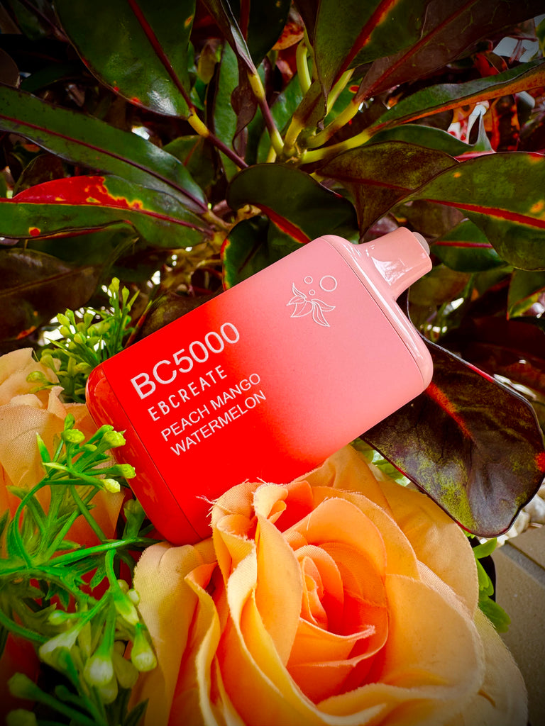 The image features a close-up of a red to peach gradient disposable vape device prominently displayed in front of a vibrant backdrop of greenery and peach-colored artificial flowers. The device is labeled "BC5000" with the flavor "Peach Mango Watermelon" indicated beneath the brand. The naturalistic setting is enhanced by the lush leaves of plants, providing a contrasting background that accentuates the bright color of the vape device. The overall composition suggests a lively and fresh outdoor ambiance.