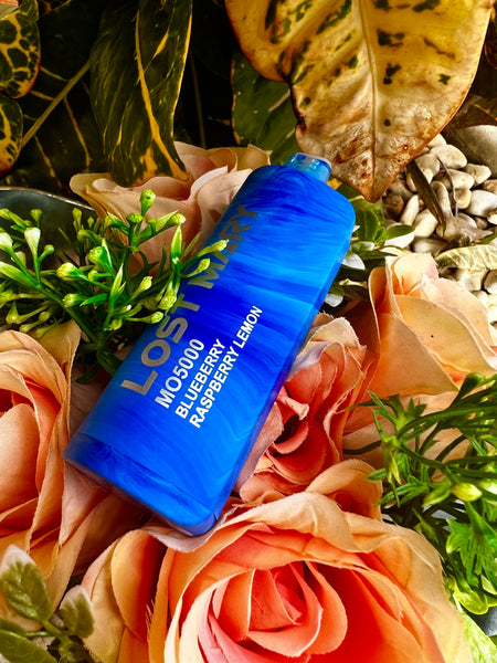 The image captures a bright blue disposable vape device with bold white text that reads "LOST MARY" and "MOS500 BLUEBERRY RASPBERRY LEMON". It is prominently positioned atop an arrangement of peach-colored artificial roses and sprigs of greenery, with variegated leaves in the background. The intense blue hue of the vape stands out vividly against the warm colors of the roses and the varied greens of the plants.