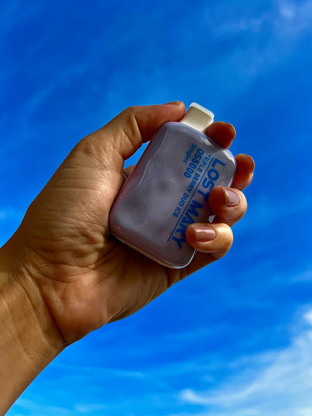 A hand is holding a white and blue Lost Mary disposable vape against a clear blue sky. The vape device has a translucent body, and the text "Lost Mary" appears in blue lettering on the side, along with the flavor description. The holder's nails are painted in a subtle pink polish, complementing the light tones of the device. The image captures a bright and airy atmosphere, with the focus on the hand presenting the vape to the expansive sky above.