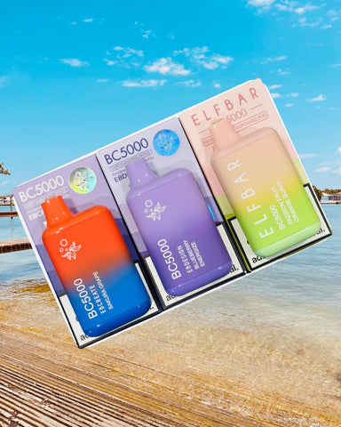 The image features three packaged vaping devices displayed in front of a coastal scene. The packages are aligned side by side. From left to right: the first package is blue with a green sticker, containing a red vaping device labeled "BC5000 EBCREATE SAKURA GRAPE"; the middle package is light purple with a blue sticker, holding a purple device reading "BC5000 EBDESIGN BLUEBERRY ENERGIZE"; and the third package is peach-colored with a green vaping device inside, marked "ELFBAR BC5000 PASSION FRUIT ORANGE GUAVA". The background shows a clear blue sky and calm sea with a wooden pier extending into the water. The presentation creates an impression of a product lineup against a tranquil, sunny seaside setting.