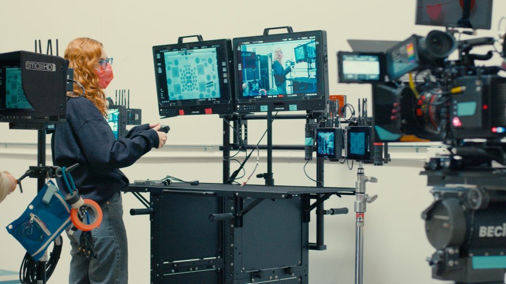 A woman uses a wireless controller to adjust the lens of a cinema camera. She's reviewing the live feed on a monitor in front of her. The monitor is one of two that are mounted to an INOVATIV Deploy.