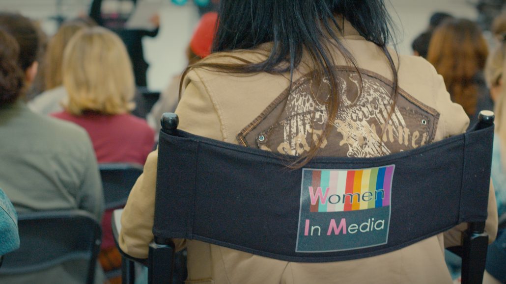 A female attendee sits in a director's chair that displays the Women in Media logo. The logo features alternative branding which includes the the Progress Pride flag colors.
