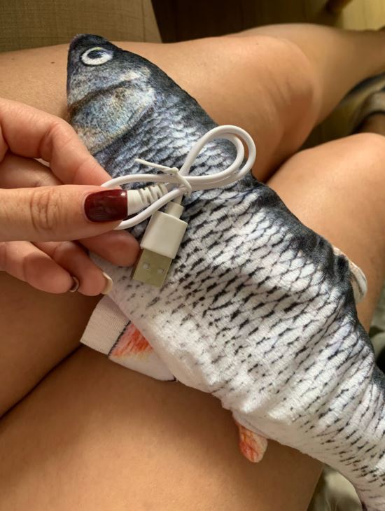Woman holding a battery-operated mechanical flopping fish cat toy on her lap, showing its charging cable for the Floppy Fish interactive cat toy