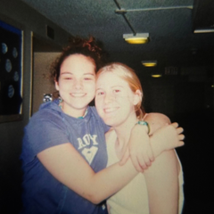 Two girls hugging. One is wearing a blue short sleeved shirt and the other is wearing a white tank top.