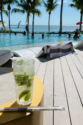 A cold coctail drink resting on a poolside deck with palm trees and ocean in the background