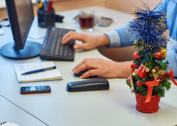 Close-up of employee’s hands as he sits at desk decorated for holiday season, miniature Christmas tree near his phone and computer.