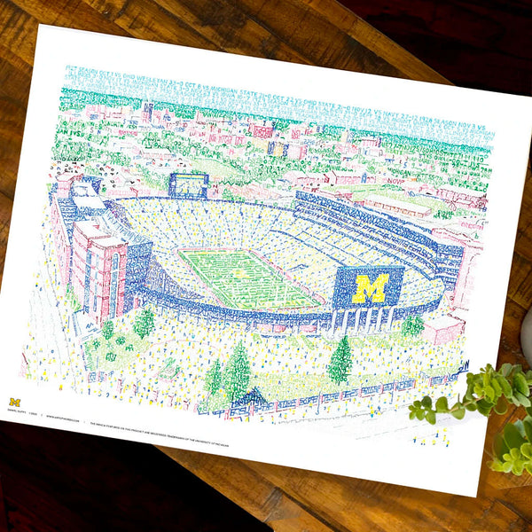 Unframed word art print depicting Michigan Stadium, handwritten with stats of 440 Wolverines wins, lies flat on table.