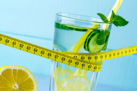 An image of a glass of water with lemon and lime slices, a straw, and a half of a lemon the side with a tape measure on a blue background.