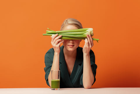 An image of a blond woman sitting at a table with a glass of celery juice while holding a stalk of celery in front of her face.