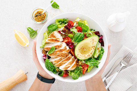 An image of hands holding a white bowl filled with grilled chicken meat and fresh vegetable salad of tomato, avocado, lettuce and spinach.