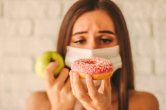An image of a young woman in protective mask on face deciding between an apple and a donut. 