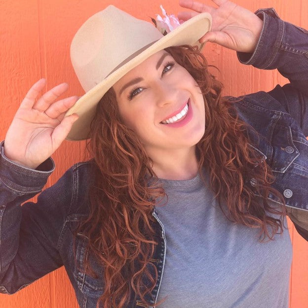 curly haired woman smiling, holding her hat on her head with both hands