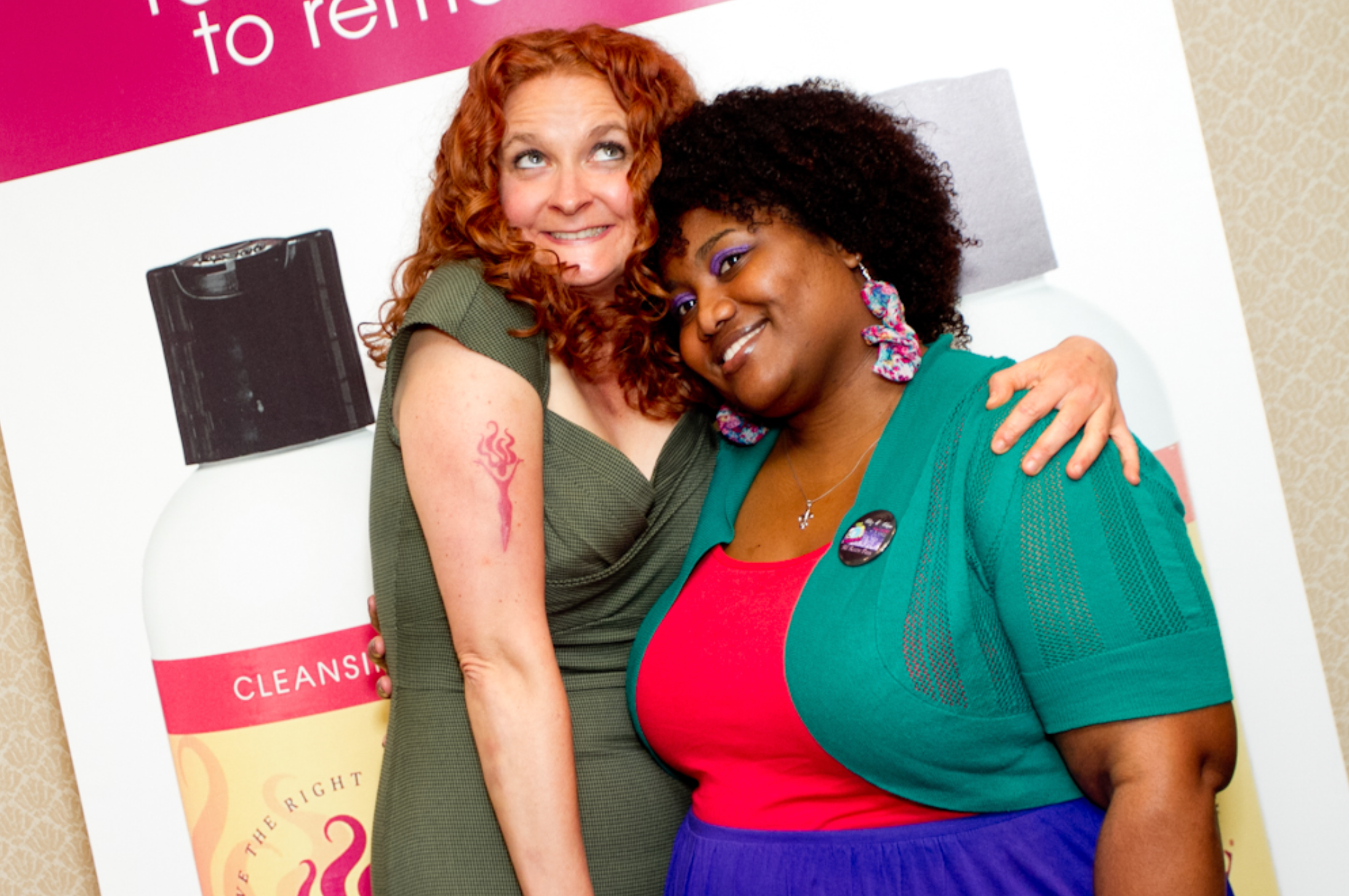 two curly haired woman hugging and smiling for the camera against a poster