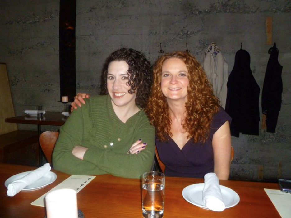 Two curly haired woman sitting at a dinner table, one with their arm around the other