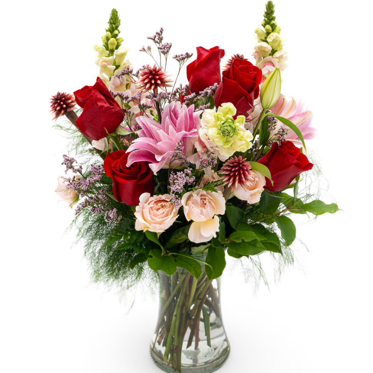 Spray roses, Double lily, snapdragon, red rose and stella shine in reds, pinks and creams. Soft whisps of green tree fern and light pink limonium spread across the arrangement.