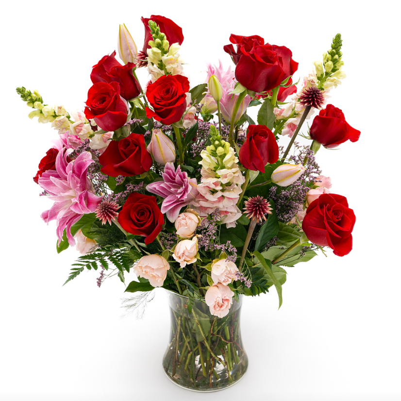 Spray roses, Double lily, snapdragon, red rose and stella shine in reds, pinks and creams. Soft whisps of green tree fern and light pink limonium spread across the arrangement.