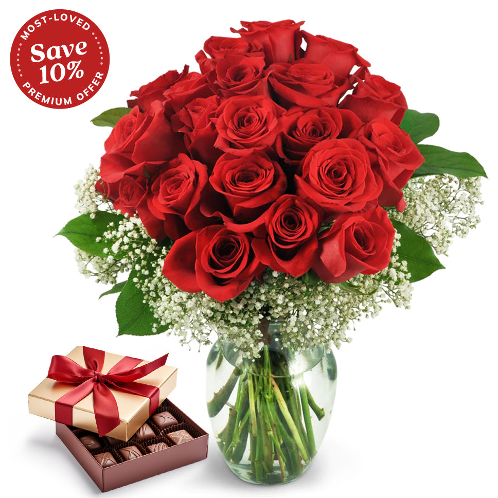 A vibrant arrangement of two dozen red roses with baby's breath in a clear glass vase paired with a gold chocolate gift box tied with a red satin ribbon, filled with assorted chocolates. Premium selection features a 10% savings.