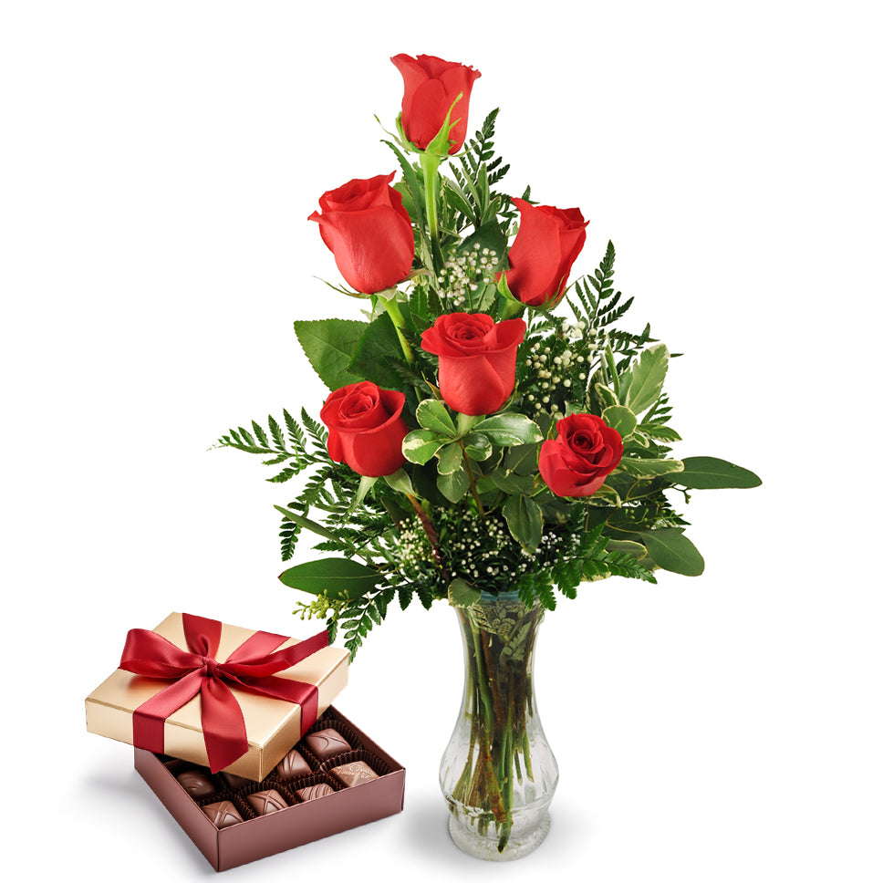 Six red roses with baby's breath in a clear glass vase paired with a gold chocolate gift box tied with a red satin ribbon, filled with assorted chocolates. 