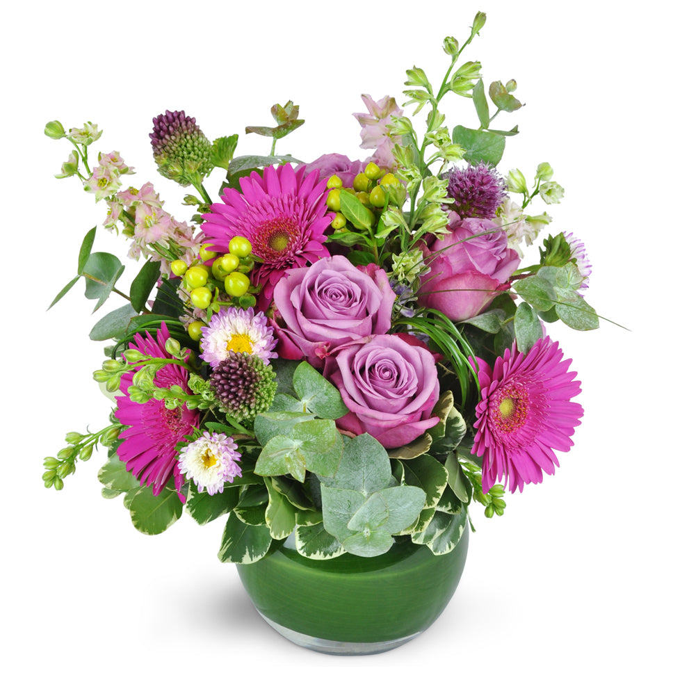 Parisian Garden. A gorgeous mix of larkspur, gerbera daisies,  lavender roses, garden roses, and bupleurum are elegantly arranged in a round glass vase.