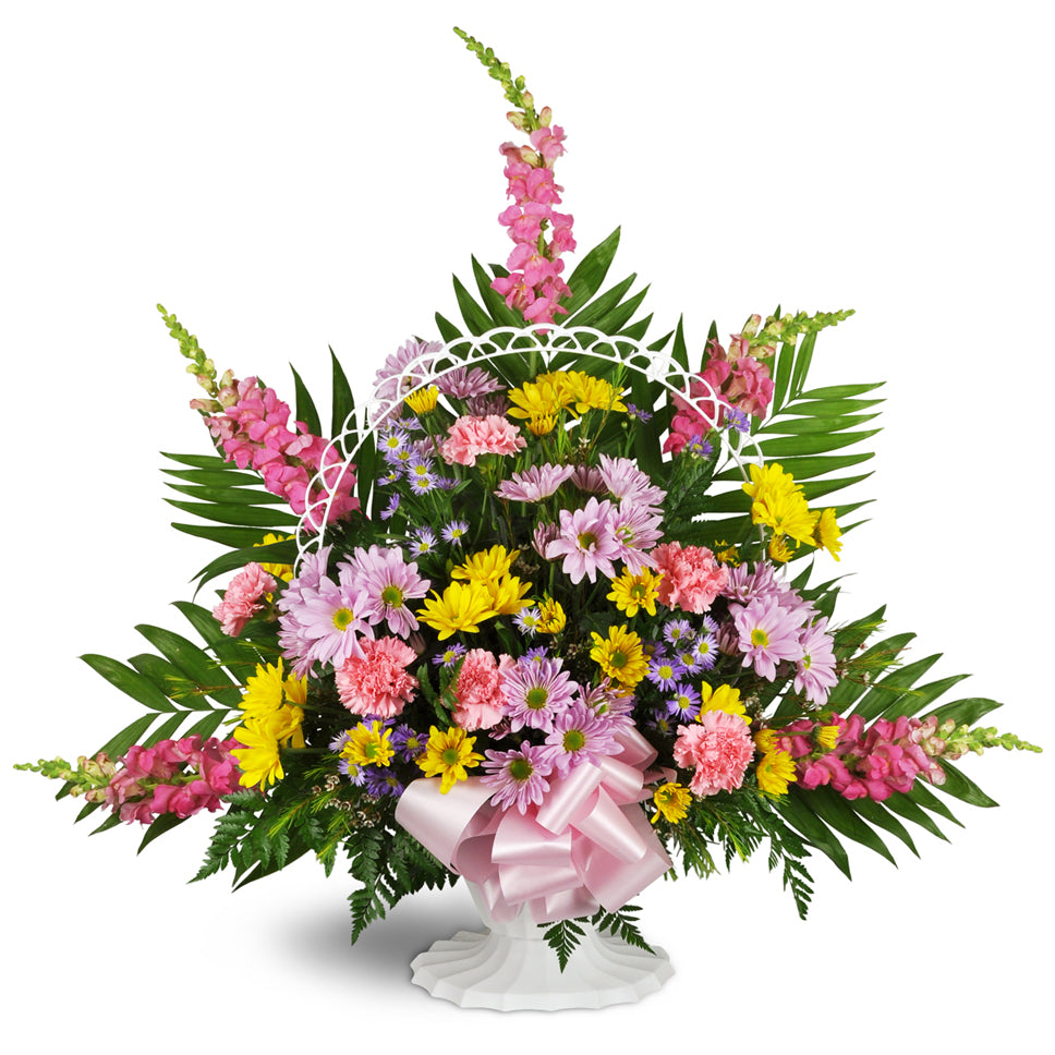 Shades of Remembrance™. Pink snapdragons, green fern fronds, yellow and purple daisies, and pink carnations are arranged in a white basket and accented by a decorative white lattice arch and pink ribbon.