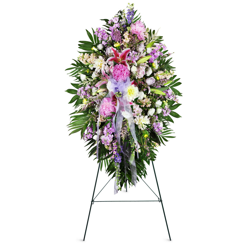 Tender Sentiments Lily Spray™. This standing spray features a comforting assortment of lilies, peonies, and dahlias in pinks, whites, and yellows with stock and fresh greenery topped off with a bow.