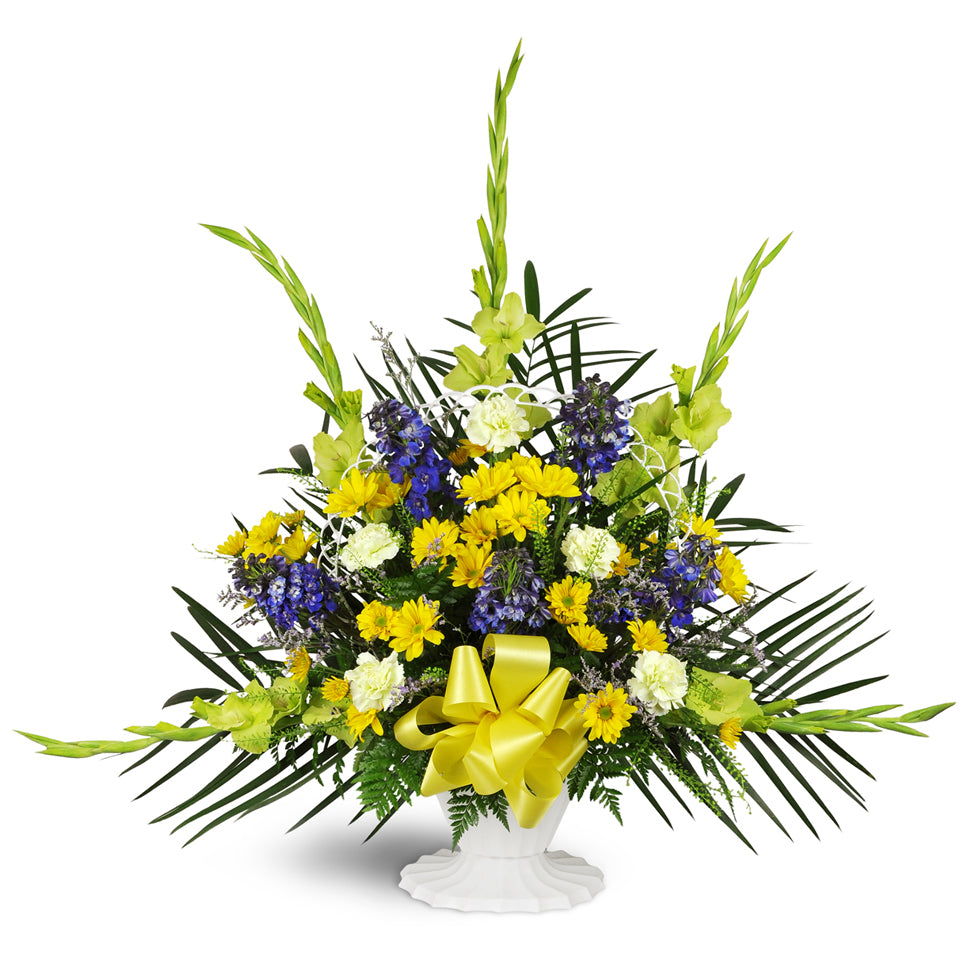 Heaven’s Promise Sympathy Tribute™ - Standard. Features a classic white basket blooming with blue delphinium, yellow daisies, and gladiolus—a yellow bow adds the finishing touch.
