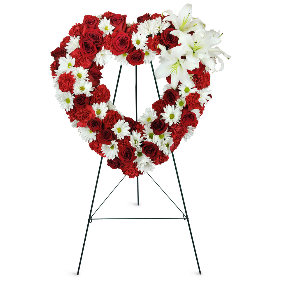 Rosy Remembrance Heart Wreath™. Red roses, white lilies, white daisies, and red carnations are arranged with greens for display at the funeral or service.