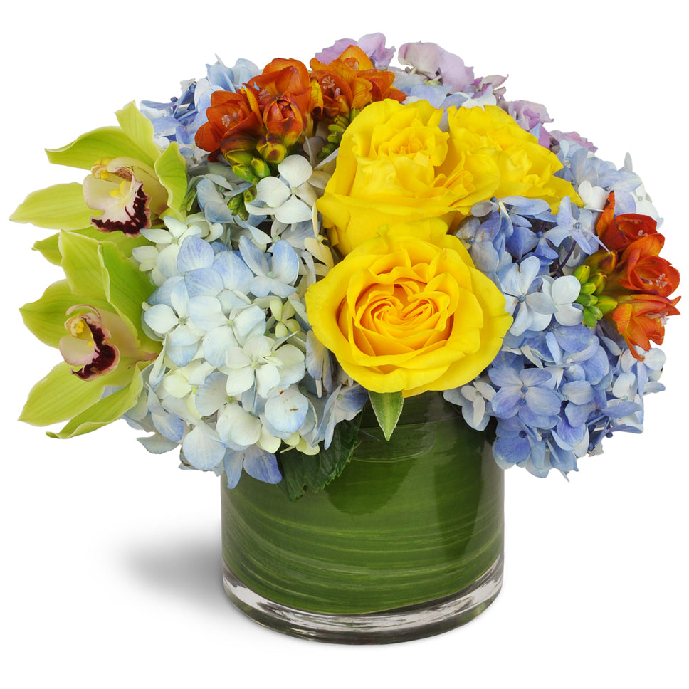 Clear Skies Bouquet™. Yellow roses, green cymbidium orchids, blue hydrangea, and orange freesias are cheerfully arranged in an aspidistra-lined clear glass vase.