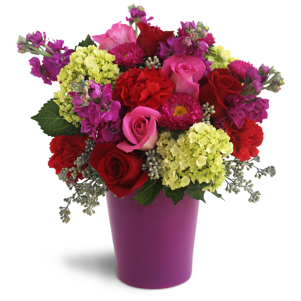 Bursting with Blooms floral arrangement. Pink roses, red roses, red carnations, purple phlox, green hydrangeas, and asters are perfectly arranged in a glass vase.
