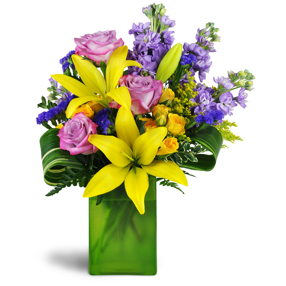 Springtime Kiss™ - Deluxe. Treat them to lavender roses, sunny yellow lilies, purple stock, and more arranged in a glass vase.