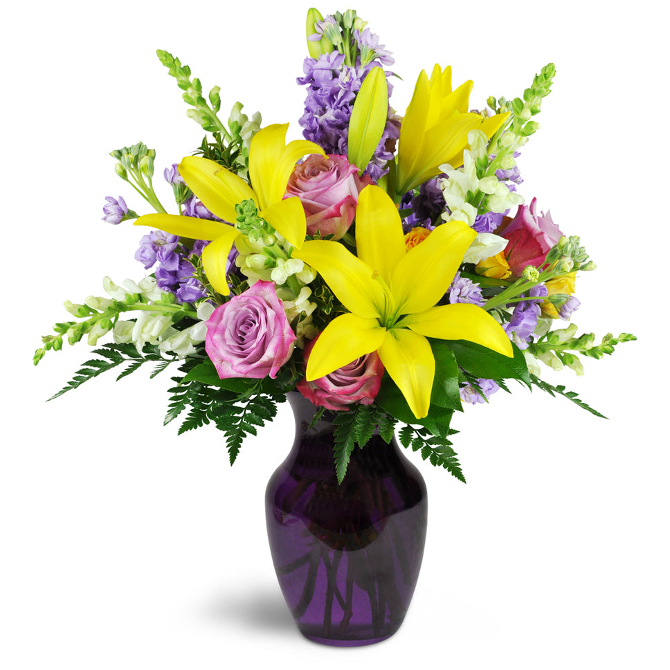 Sprightly Spring™. Yellow Asiatic lilies bloom bright with lavender roses, stock, and creamy white snapdragons in a glass vase.