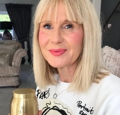 Blonde woman smiling, holding a gold urn, with a white shirt featuring black text and drawing.